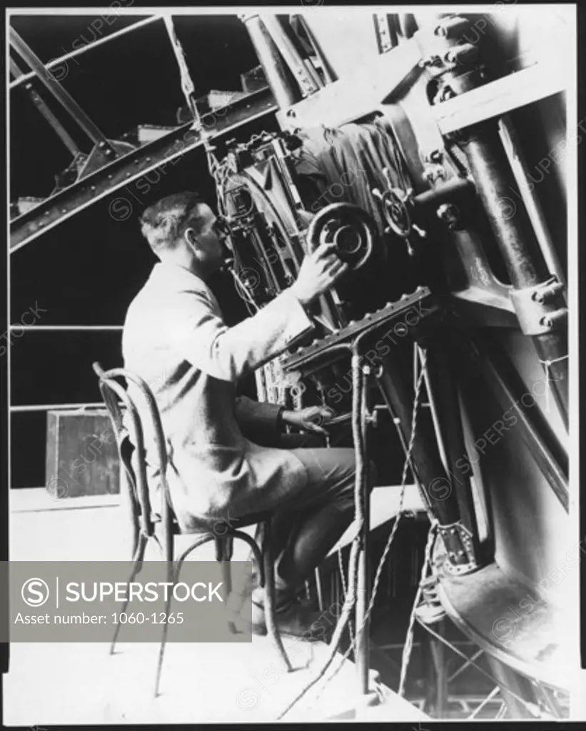 EDWIN HUBBLE SEATED IN CHAIR, LOOKING THROUGH THE NEWTONIAN FOCUS OF THE 100-INCH TELESCOPE.