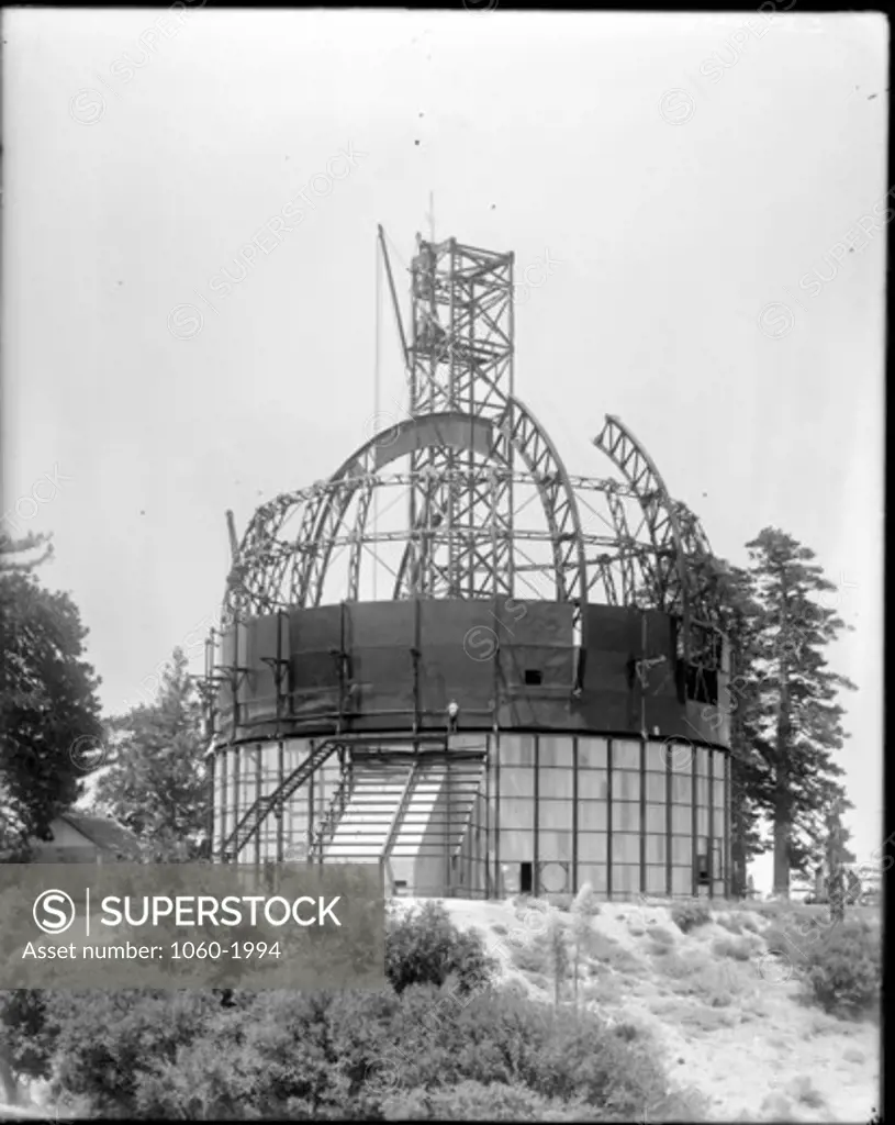 100-INCH TELESCOPE DOME BEING ERECTED WITH CRANE BASE EXTENDING FROM INSIDE THE BUILDING TO WELL ABOVE THE TOP OF THE DOME; CRANE HOISTING MAIN DOME GIRDER IN PLACE.