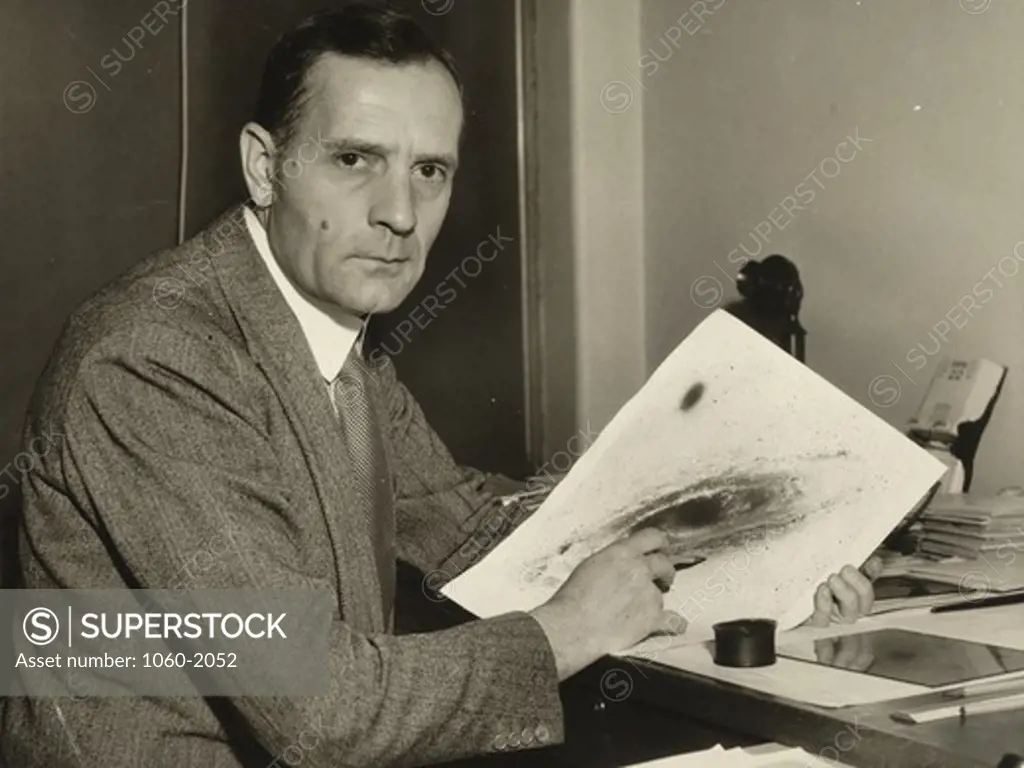 Edwin Powell Hubble at desk, holding photograph of galaxy and looking at camera