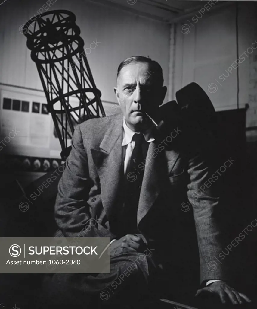 Edwin Powell Hubble sitting on desk, with astronomy exhibit behind him, including model of 100-inch Hooker, probably at Santa Barbara Street Observatory Offices
