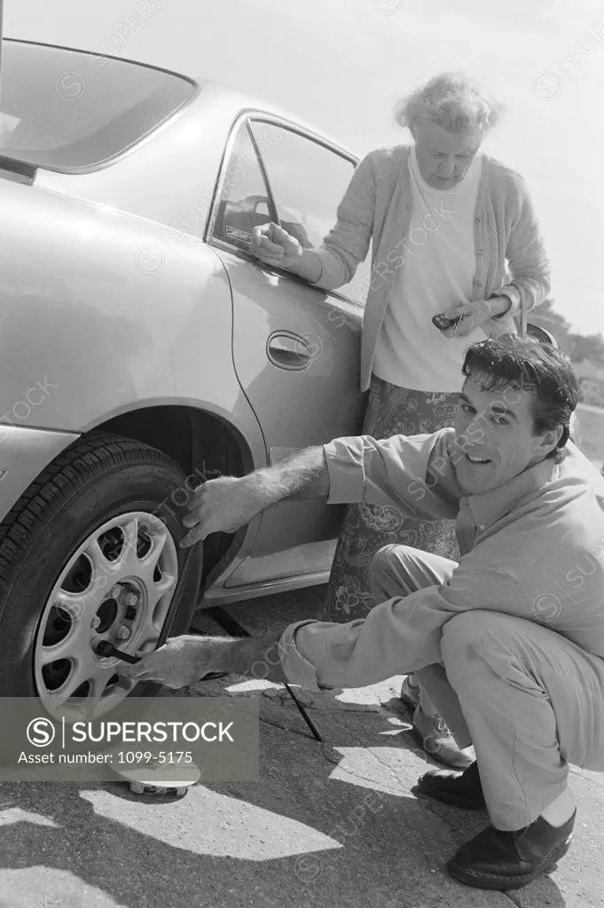 Senior woman standing beside her car with a mid adult man changing a flat tire