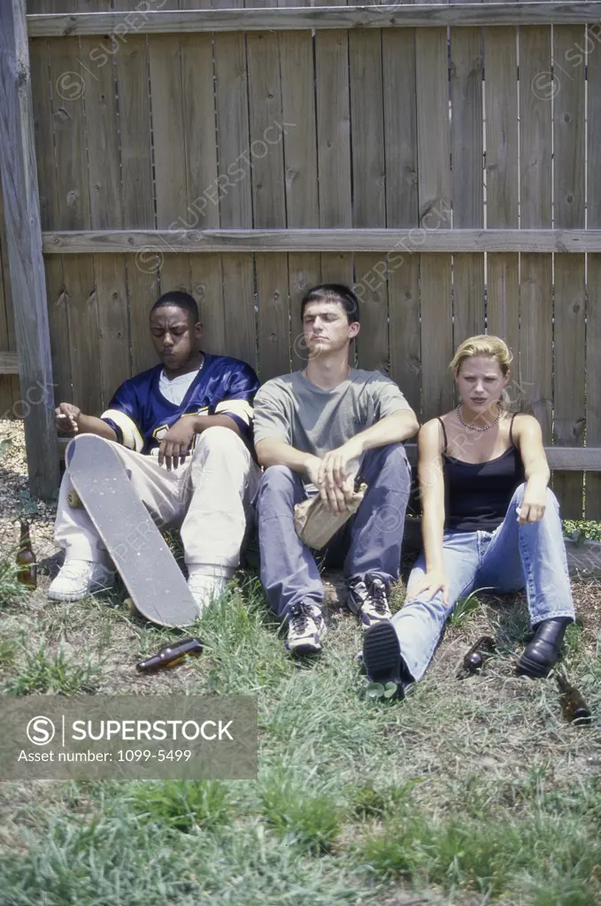 Two teenage boys and a teenage girl sitting against a fence smoking and drinking