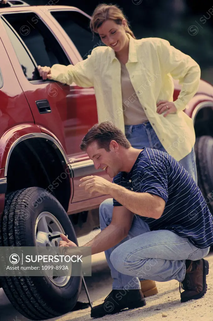 Mid adult woman standing beside her car with a mid adult man changing a flat tire