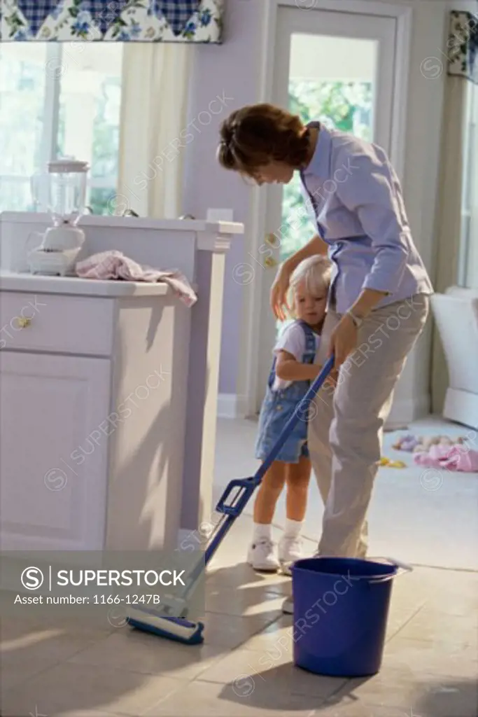 Mother cleaning the kitchen floor with her daughter standing beside her