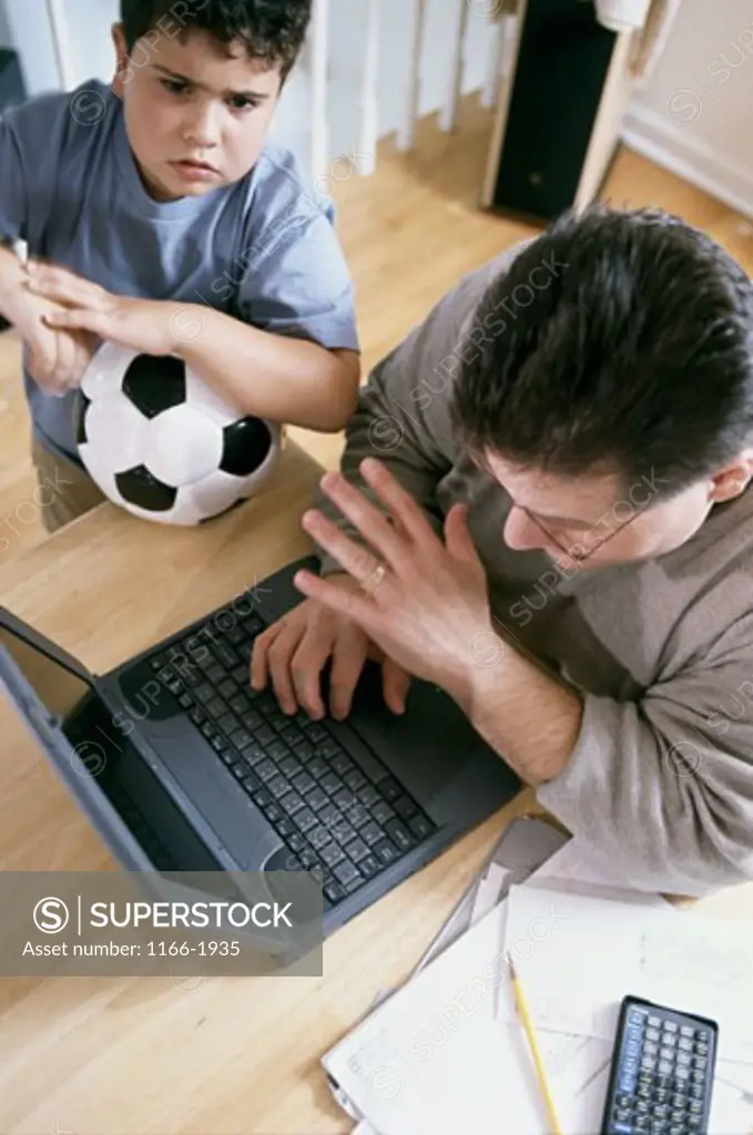 High angle view of a mid adult man working on a laptop with his son standing beside him with a soccer ball