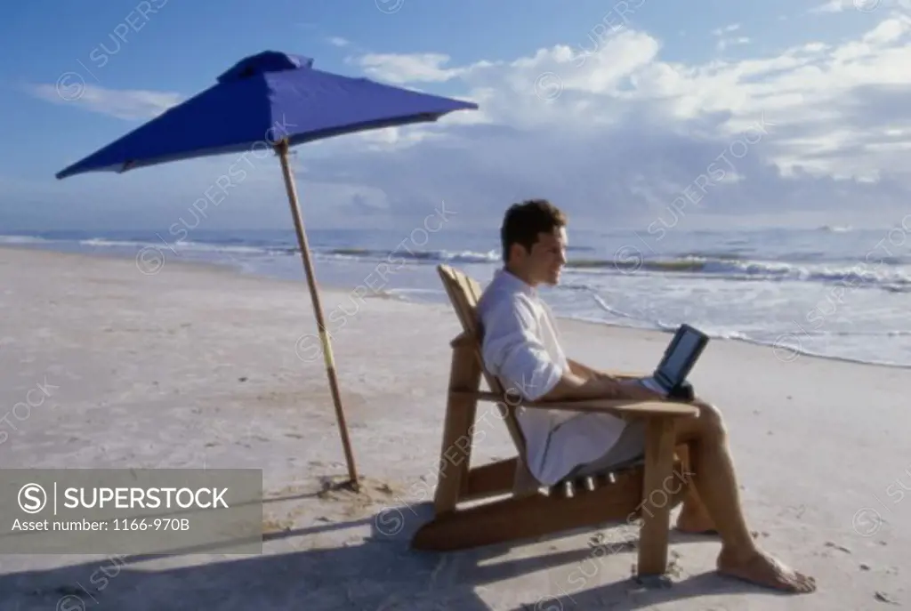 Side profile of a young man sitting on a deck chair on the beach using a laptop