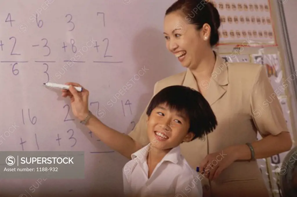 Teacher writing on a board in a classroom and a boy standing beside her