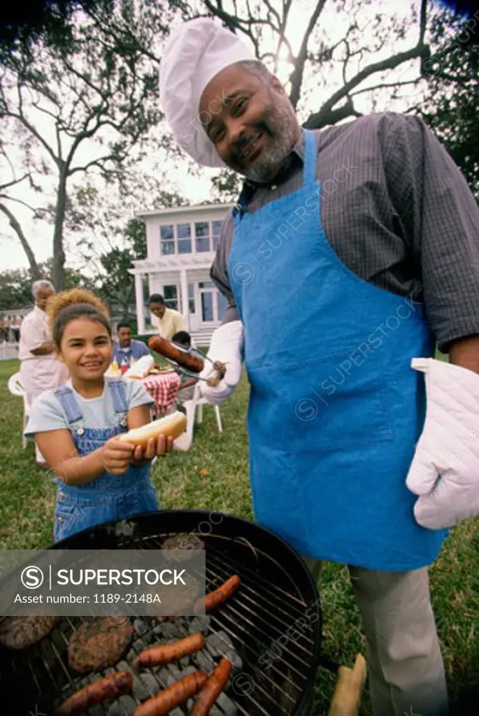 Portrait of a grandfather barbecuing hot dogs with his granddaughter standing beside him
