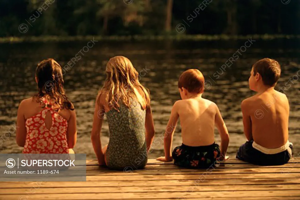 Rear view of two boys and two girls sitting side by side on a pier
