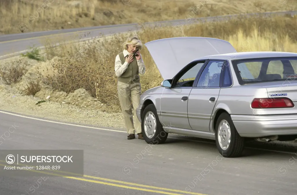 Senior woman standing in front of a broken down car talking on a mobile phone