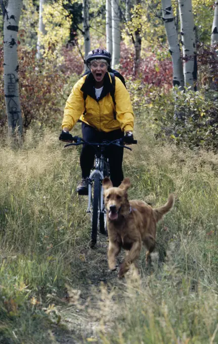 Mature woman riding mountain bike in forest and dog running in front of her