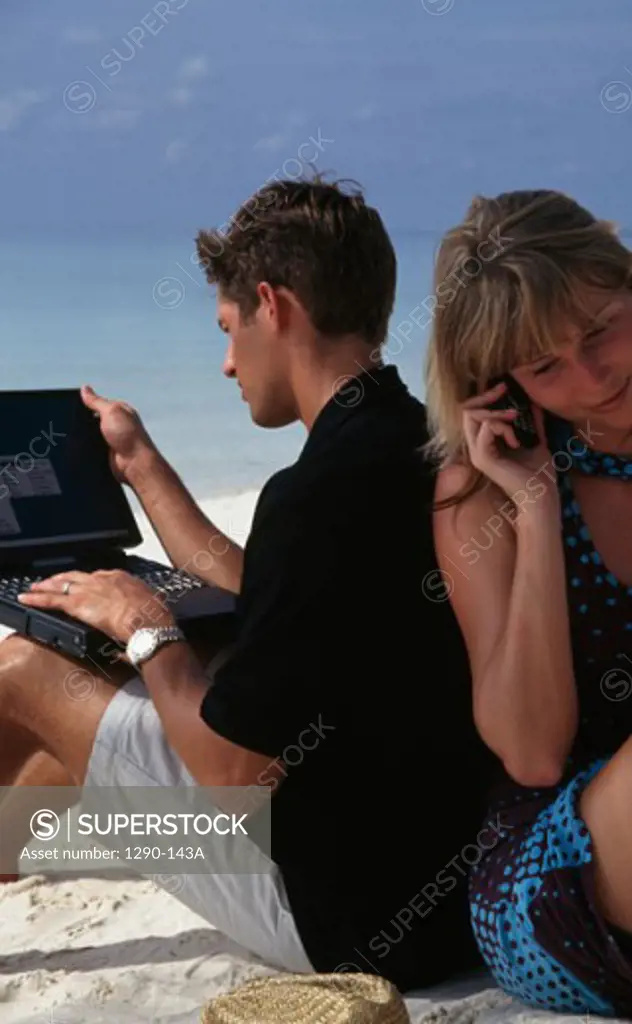 Side profile of a young man sitting on the beach using a laptop with a young woman talking on a mobile phone