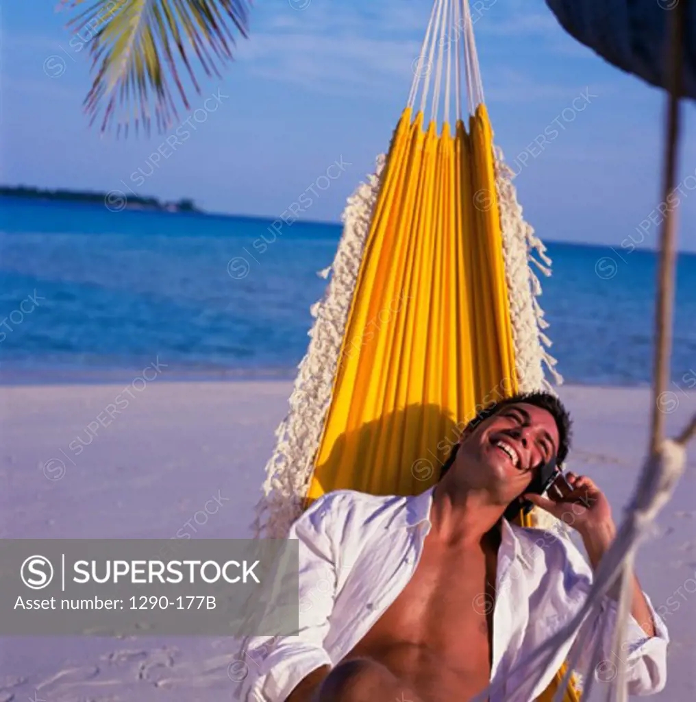 Young man lying in a hammock on the beach and talking on a mobile phone, Maldives