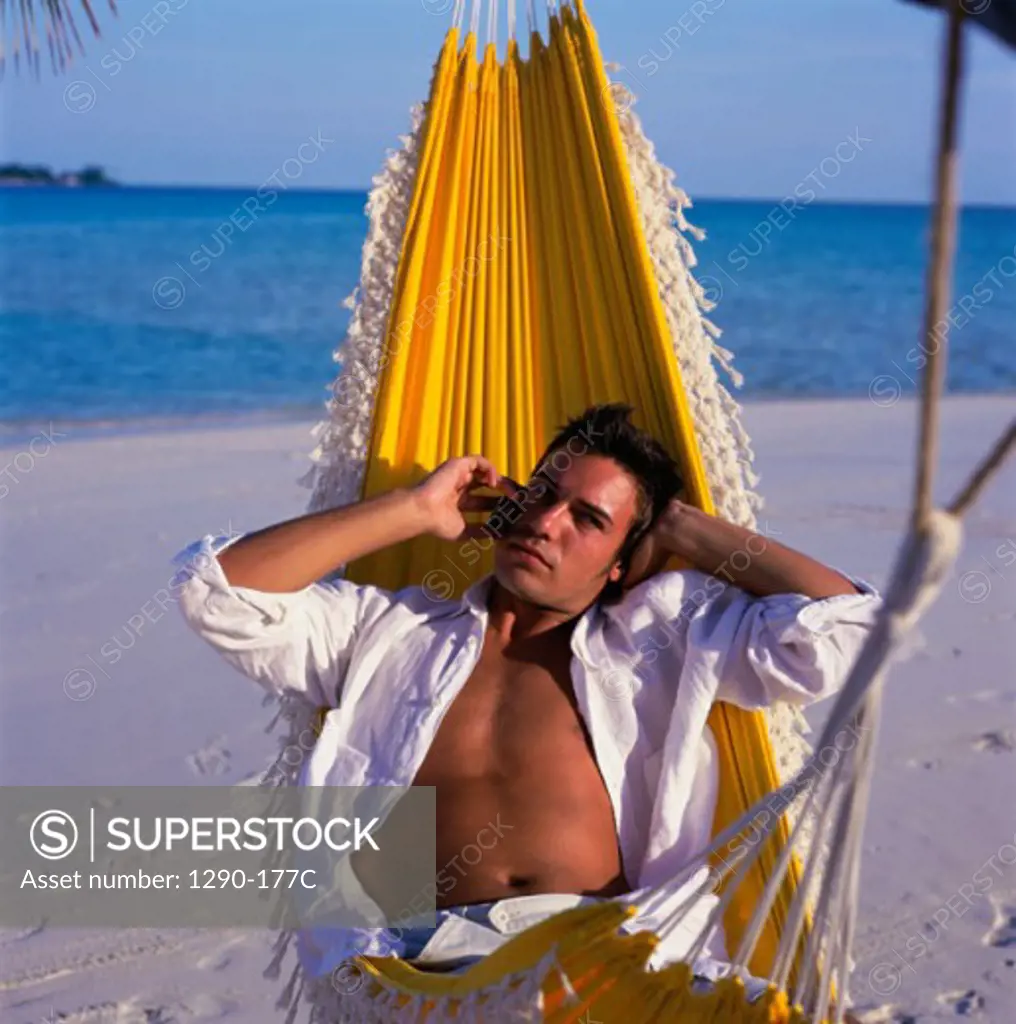 Portrait of a young man lying in a hammock on the beach talking on a mobile phone, Maldives