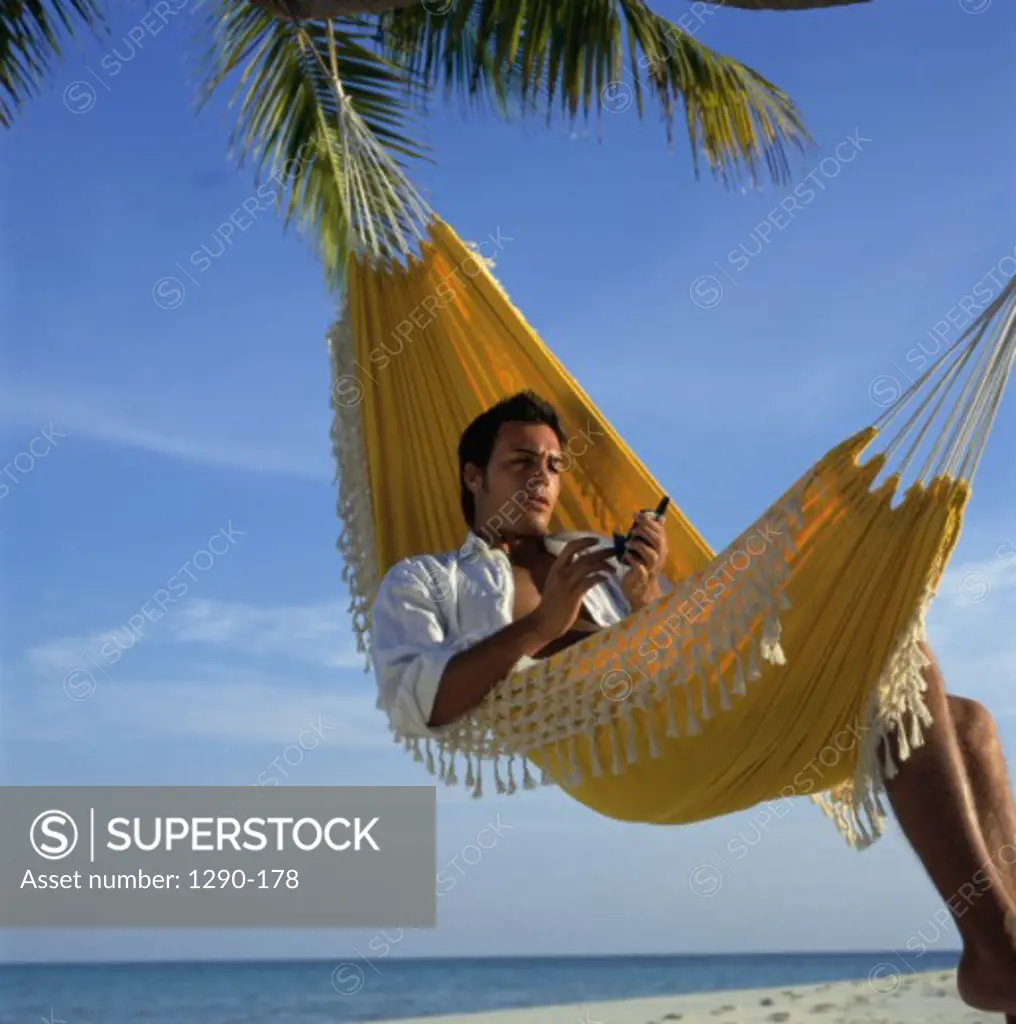 Young man lying in a hammock on the beach holding a mobile phone, Maldives
