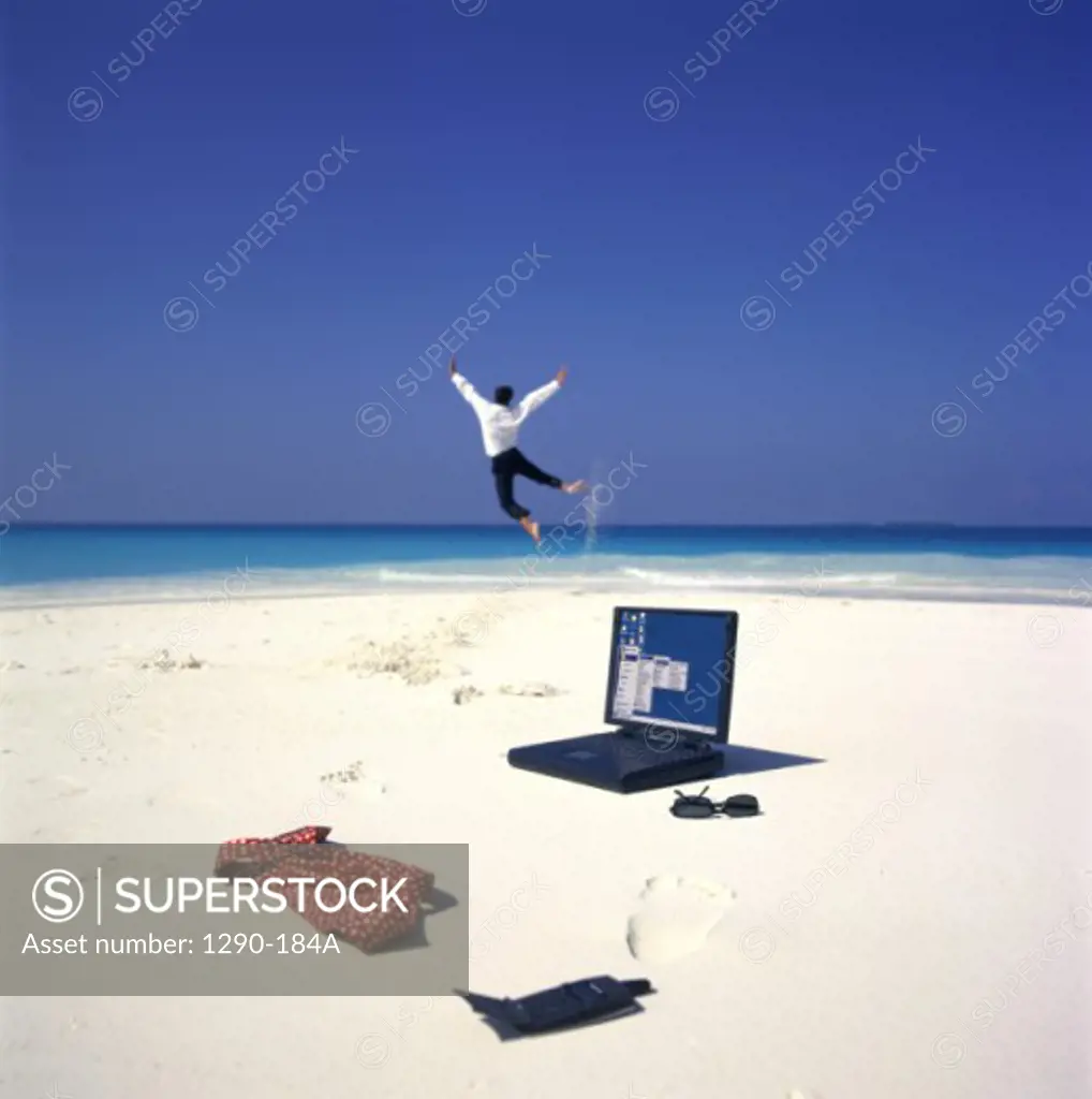 Laptop and a mobile phone on the beach with a businessman jumping in the background