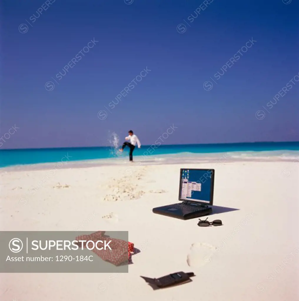 Laptop and a mobile phone on the beach with a businessman playing in water in the background