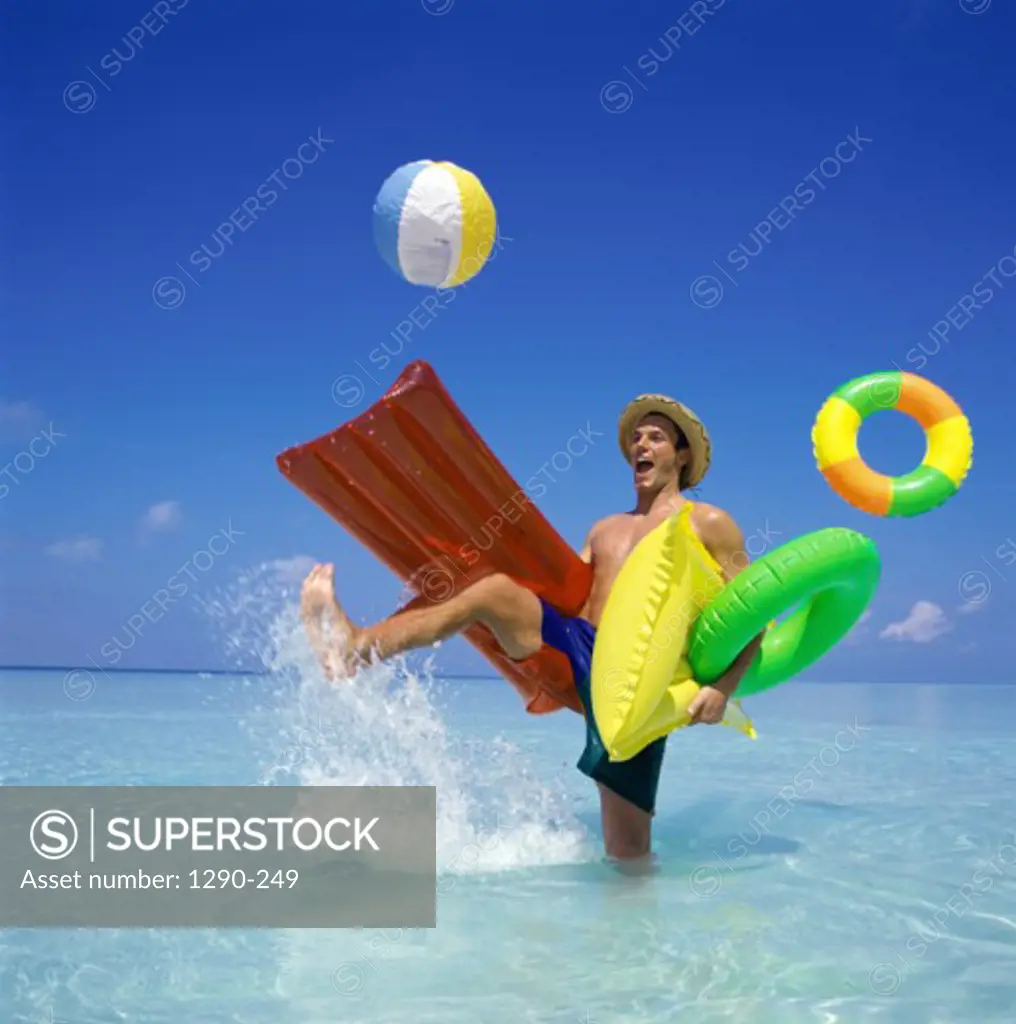 Young man standing in water holding an inflatable raft with an inflatable ring and kicking a beach ball