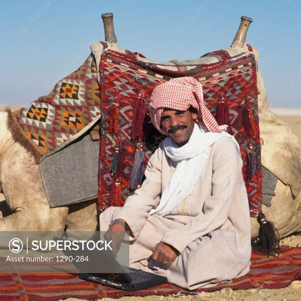 Portrait of a Bedouin man sitting in front of a laptop near a camel, Egypt