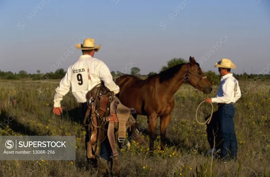 Two cowboys standing beside a horse holding a lasso and a saddle
