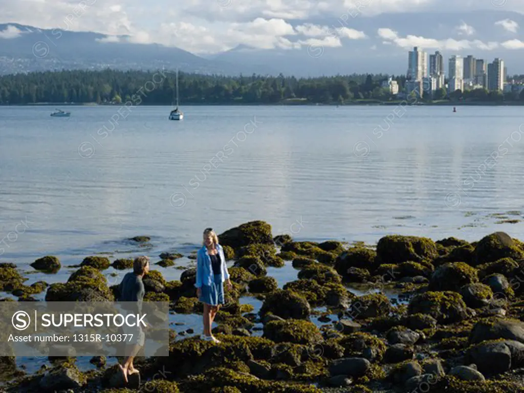 Canada, British Columbia, Vancouver, Couple walking through algae covered tidal rocks, downtown skyline in background