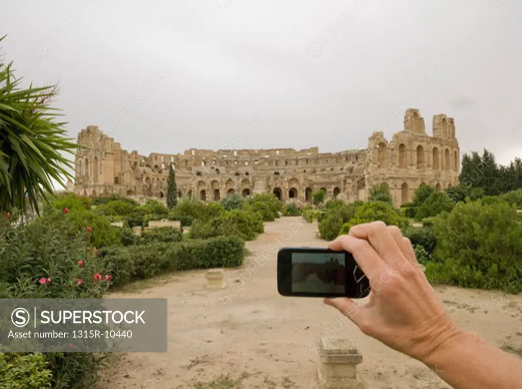Tunisia, El Djem, Woman's hand taking picture of distant coliseum with cell phone