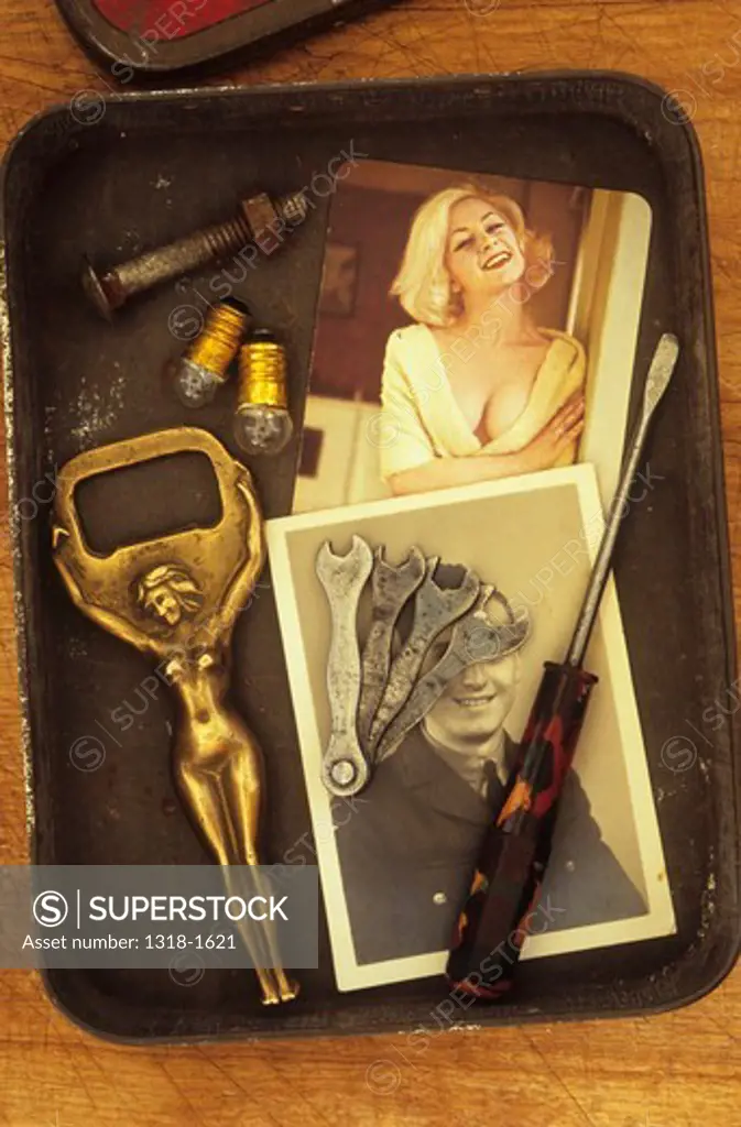 Close up of small tin containing brass naked woman bottle opener colored photo of pin-up girl black and white photo of man in armed forces uniform small screwdriver set of wrenches two flashlight bulbs and nut and bolt