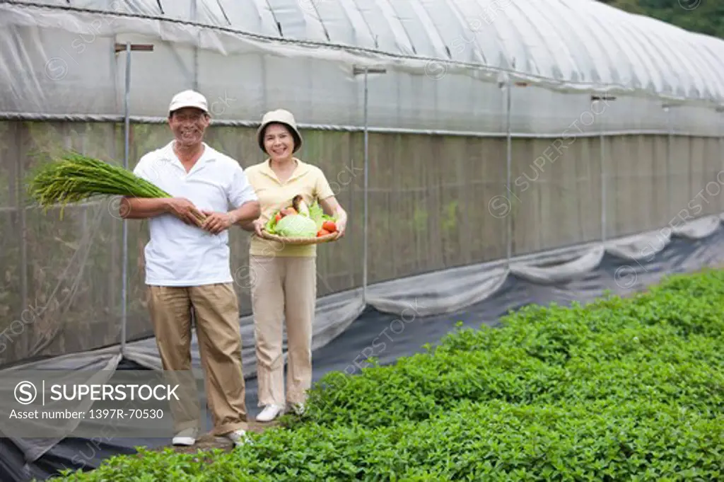 Farmer couple standing outside greenhouse with vegetables in hands