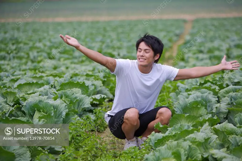 Young man crouching in the vegetable garden with arms outstretched and looking away with smile
