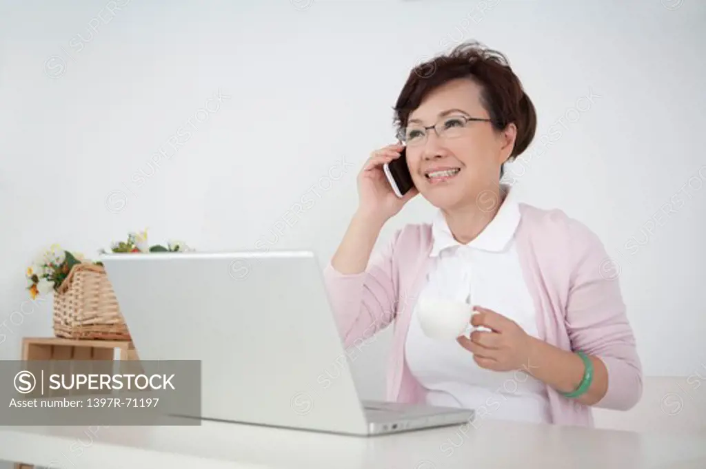 Senior woman sitting in front of a laptop and talking on cell phone