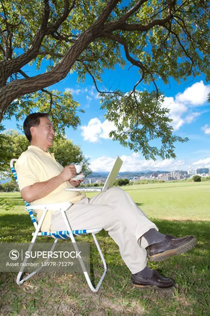 Senior man sitting in deck chair with laptop and coffee in a park
