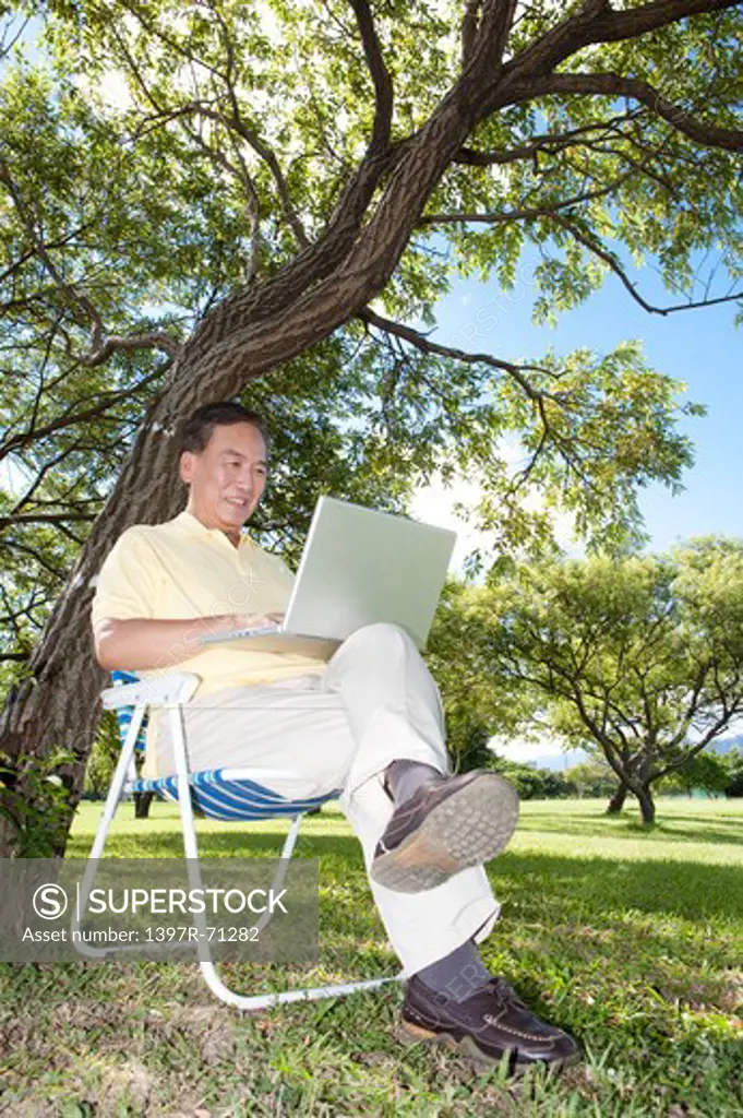 Senior man sitting in deck chair with laptop under a tree