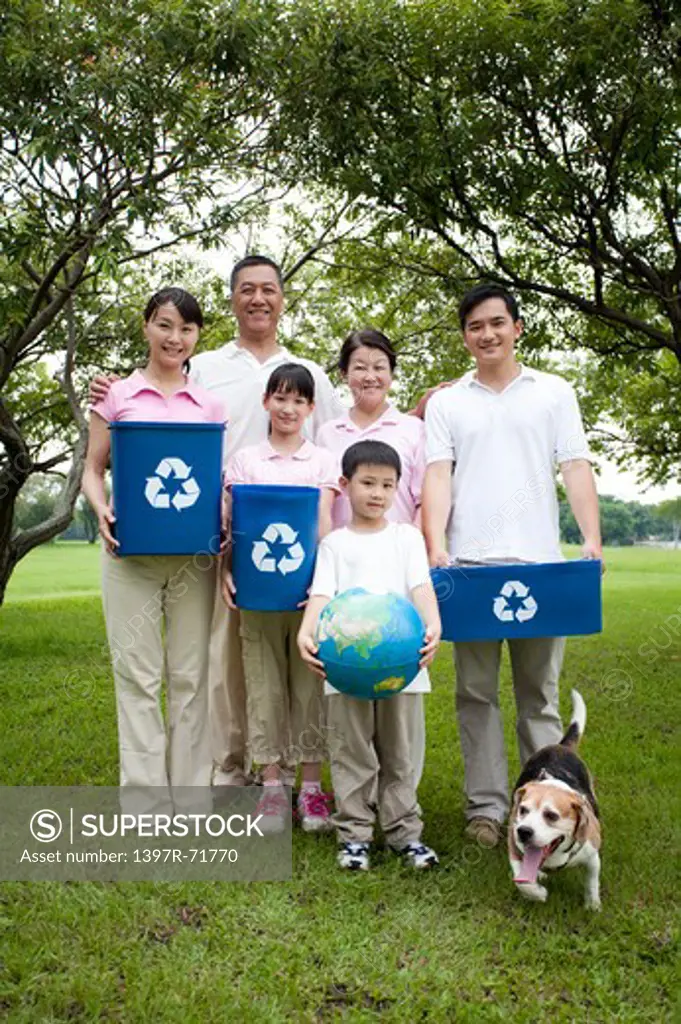 Multi-generational family standing together in a park with their dog, holding recycling bins and globe in their hands