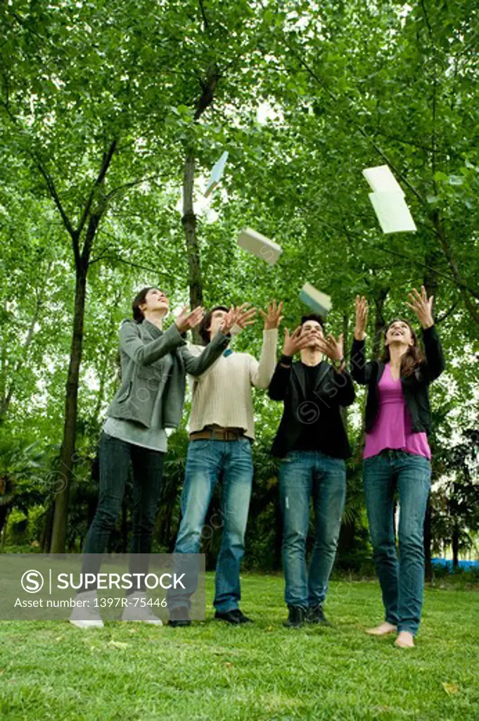 Four young friends throwing books into mid air in park