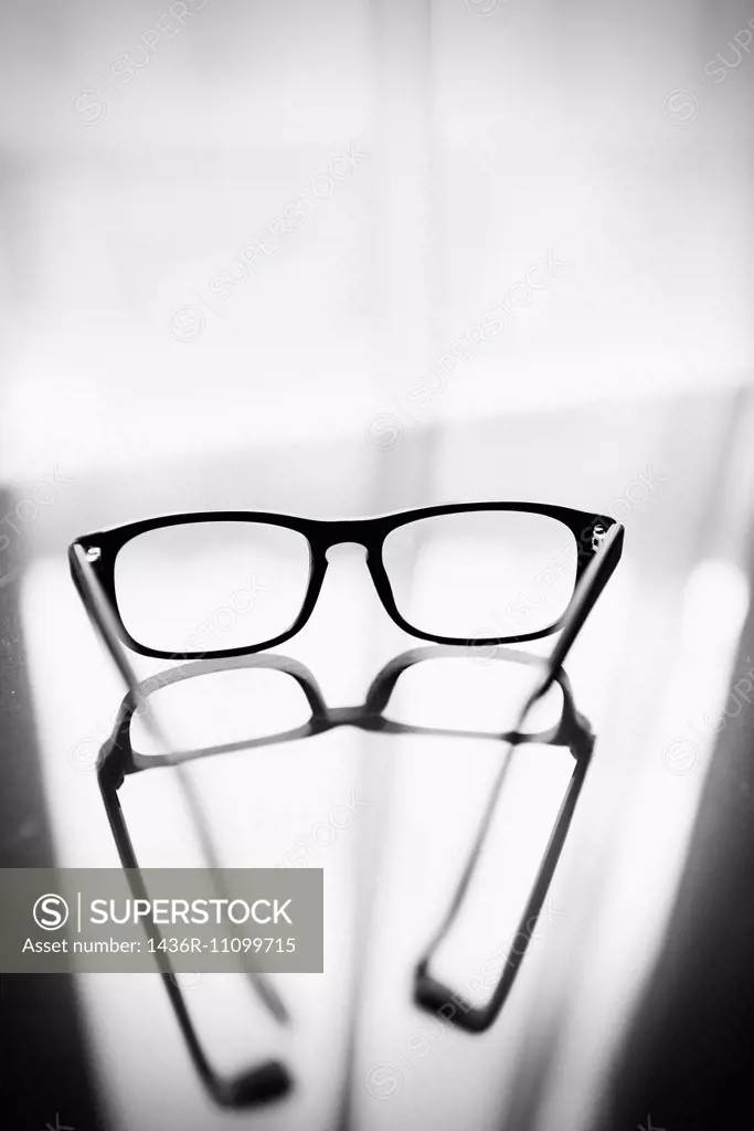 Eyeglasses on glass table in front of window with reflection of the glasses on the surface.