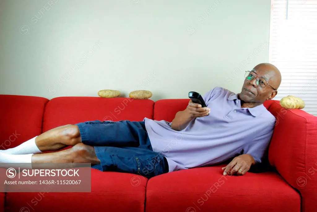 A senior citizen relaxing on a couch with a remote control in hand and potatoes on the couch.
