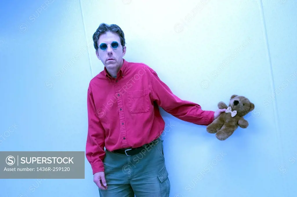 Middle-age man, wearing sunglasses and standing by a wall, holding a stuffed teddy bear