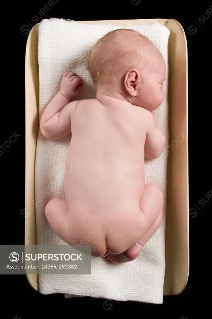 A fair-haired and naked newborn baby lies on a white blanket atop a vintage baby scale; black background.