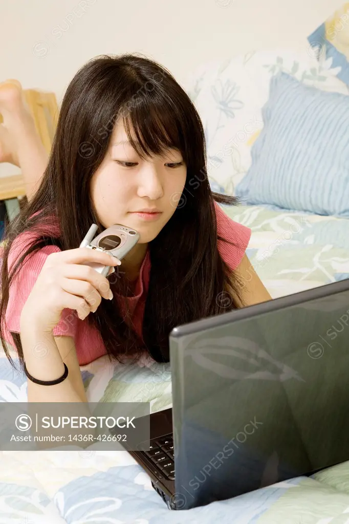 Asian American teen working on laptop computer and talking on cell phone