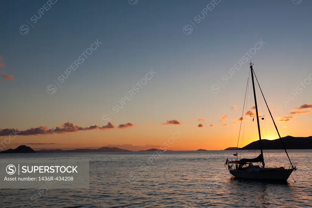 Silhouette of sailboat moored in water with orange glow of sunset lighting up the clouds and sky in Caribbean