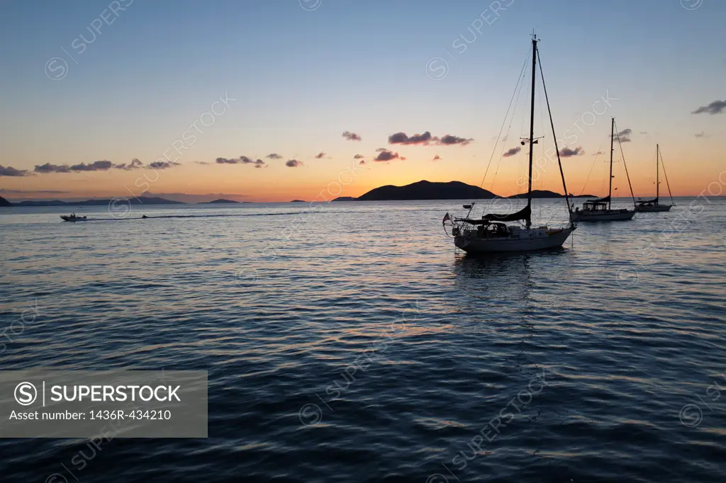 Silhouette of sailboats moored in water and power boat riding by in the orange glow of sunset in the Caribbean