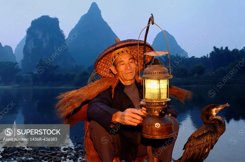 Cormorant fisherman with lamp and bird on bamboo raft at dawn on the shore of the Li river Yangshuo China