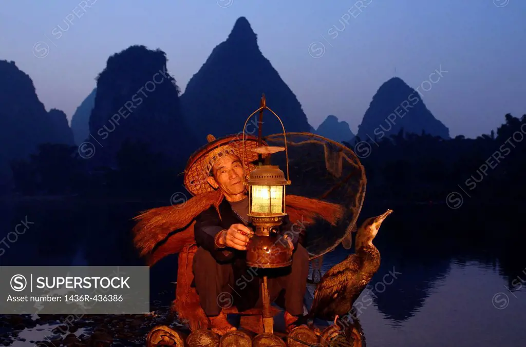 Cormorant fisherman lighting lamp with bird on bamboo raft at dawn on the shore of the Li river Yangshuo China