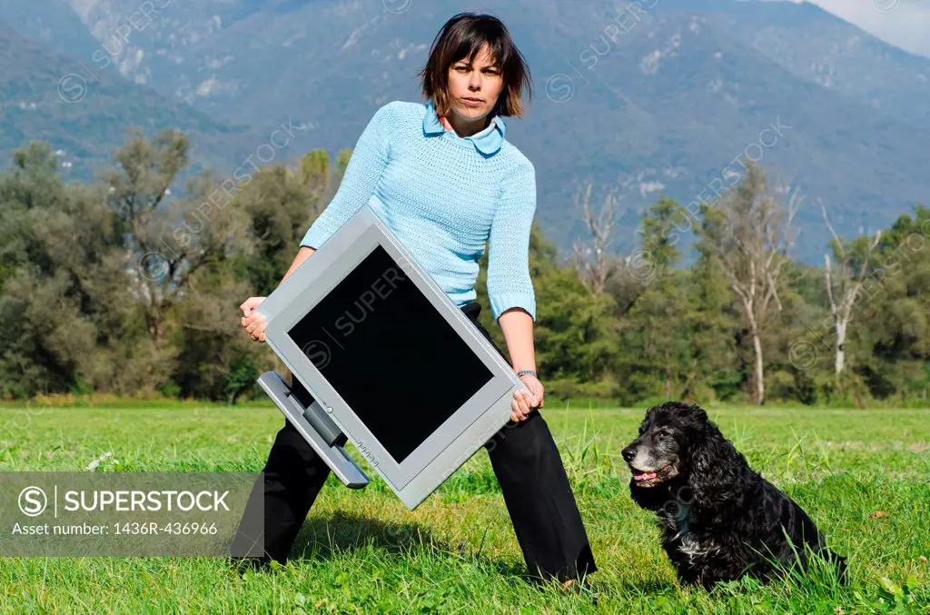 Elegant woman standing up on the field and holding a television and she have a dog