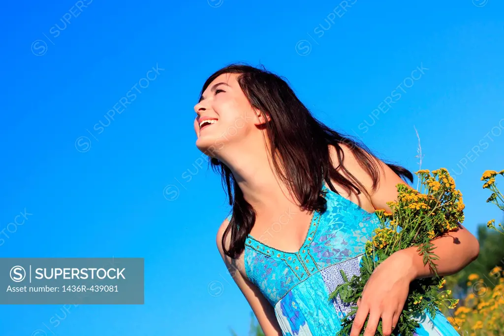 Young, beautiful girl in a blue dress holding wildflowers and looking up with a smile