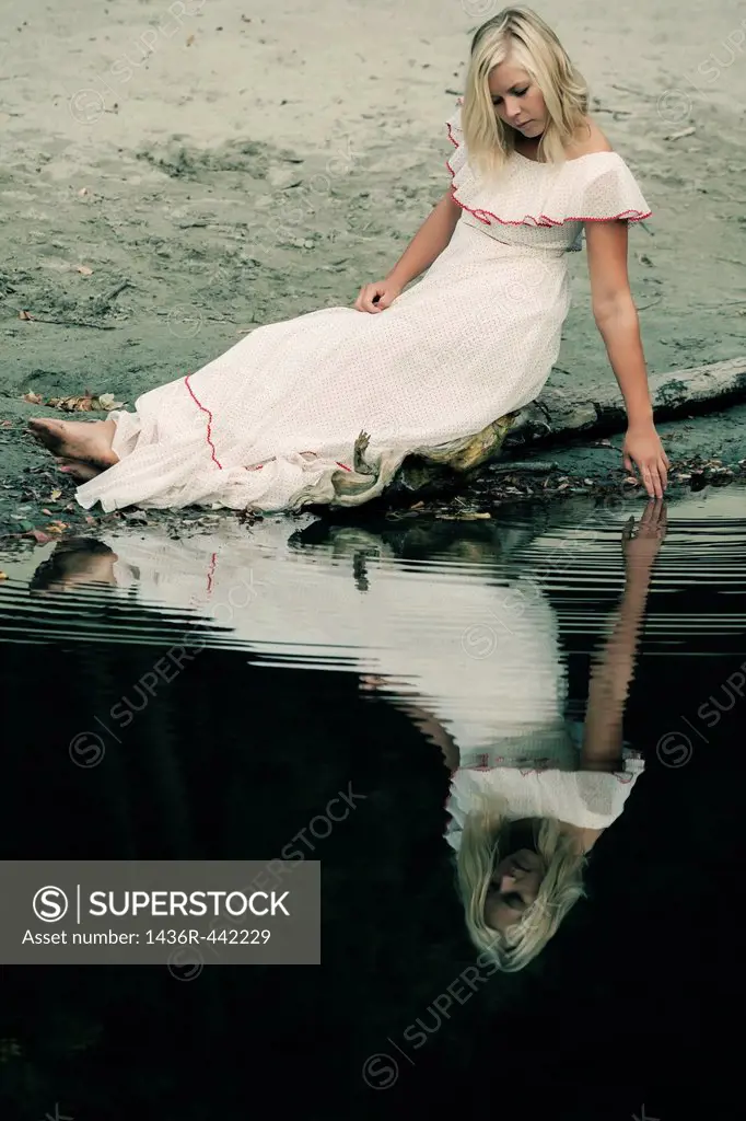 a woman in a white dress is sitting on a trunk near a pond and playing with her fingers in the water