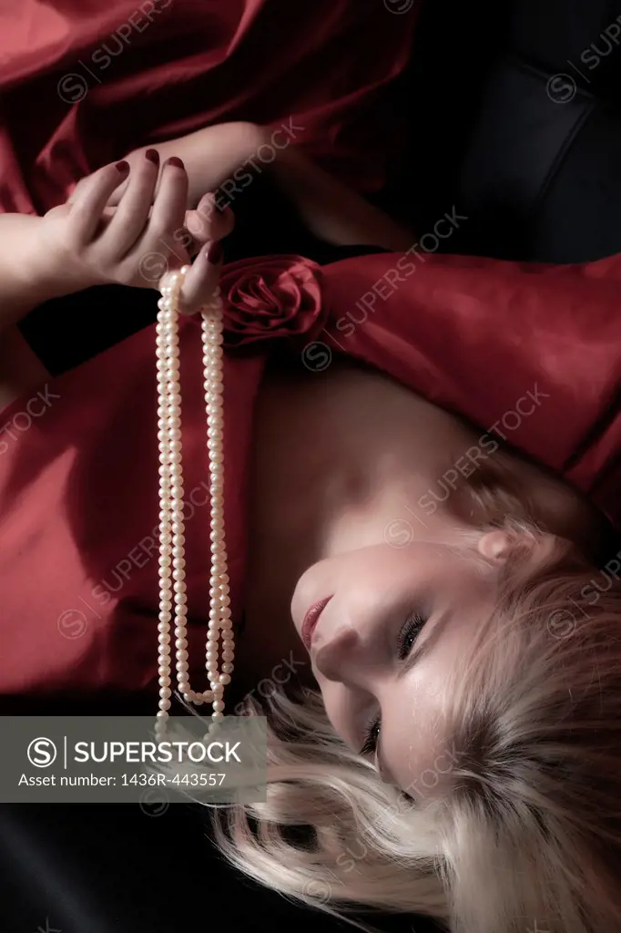 a blond woman is lying on a black sofa, holding a pearl necklace