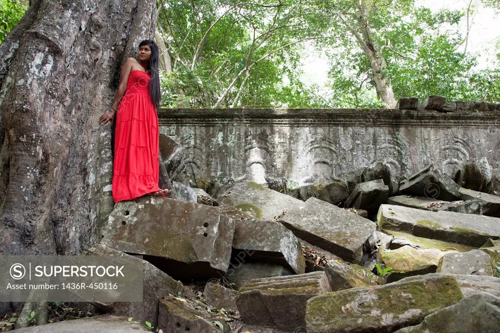 Beautiful Cambodian woman wearing a red dress posing beside a tree in Ta Phrom temple in Angkor Cambodia.