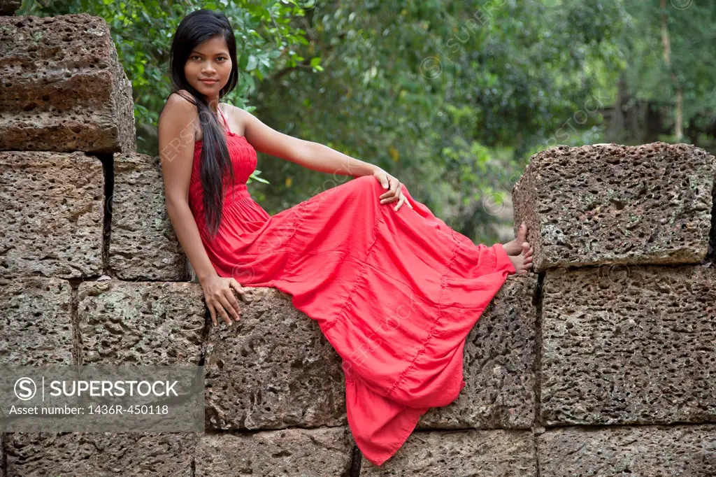 Beautiful Cambodian woman wearing a red dress sitting on a wall in Ta Phrom temple in Angkor Cambodia.