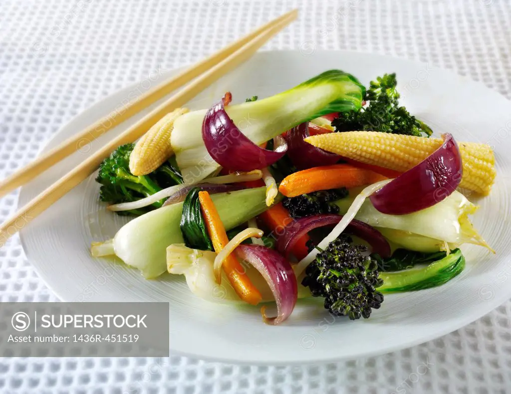 Oriental vegetarian stir fry being lifted by chop sticks in a table setting with rice & a chilli dipping sauce.